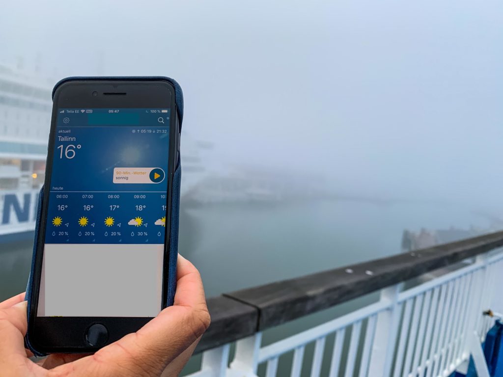 Eine Hand hält ein Smartphone ins Bild. Auf der App ist die Wettervorhersage zu sehen. Es ist Sonne und 16 Grad vorhergesagt, draußen sieht man kaum etwas uns es ist sehr sehr neblig.