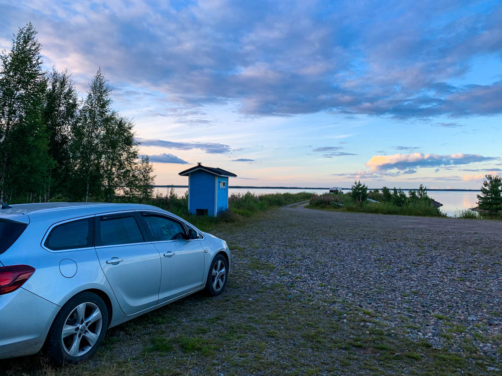 Der Mini-Camper steht an einem See in Finnland, im Hintergrund ist ein blaues Häuschen zu sehen. der Himmel ist in Sonnenuntergangsstimmung getaucht.