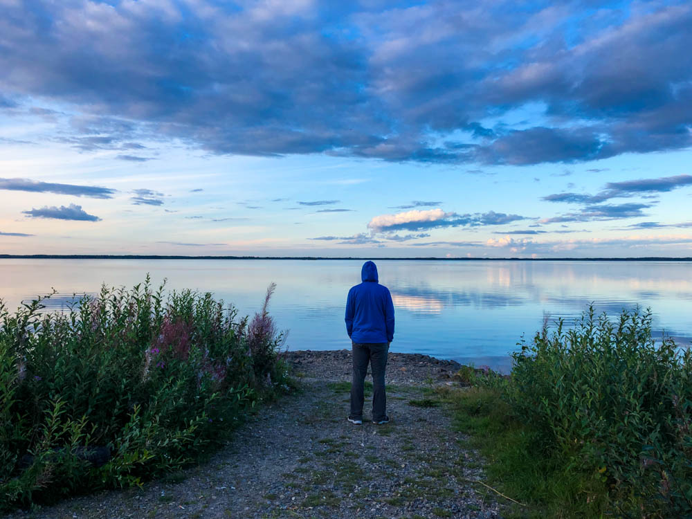 Julian steht am See kurz vor Sonnenuntergang. Die Wolken beginnen sich leicht zu färben. Von der Küste ins Landesinnere Finnlands und zurück