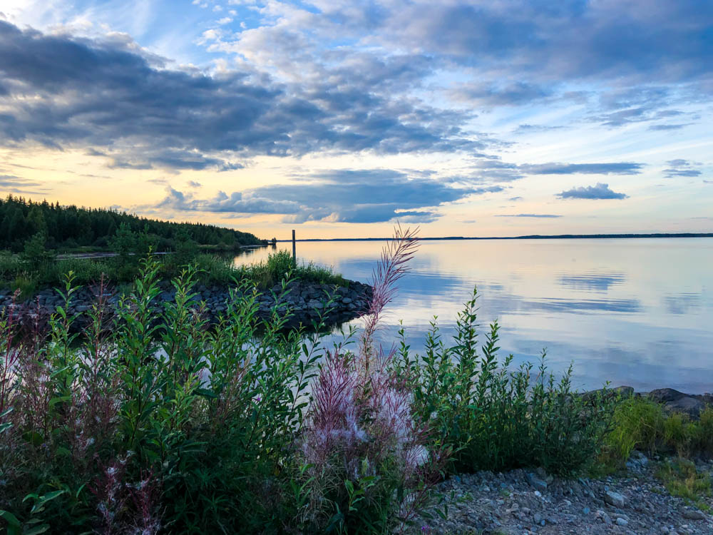 Seeufer mit einigen Pflanzen. Der Himmel über dem See spiegelt Sonnenuntergangsstimmung wieder. Von der Küste ins Landesinnere Finnlands und zurück