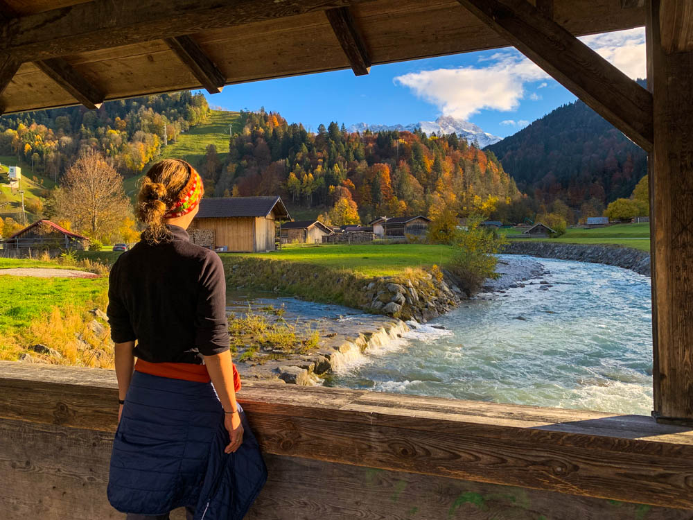 Deutsche Alpen - Melanie steht auf einer Brücke, welche über die Partnach führt. Blick auf Partnach, Heuhütten, herbstlicher Wald und verschneite Berge.