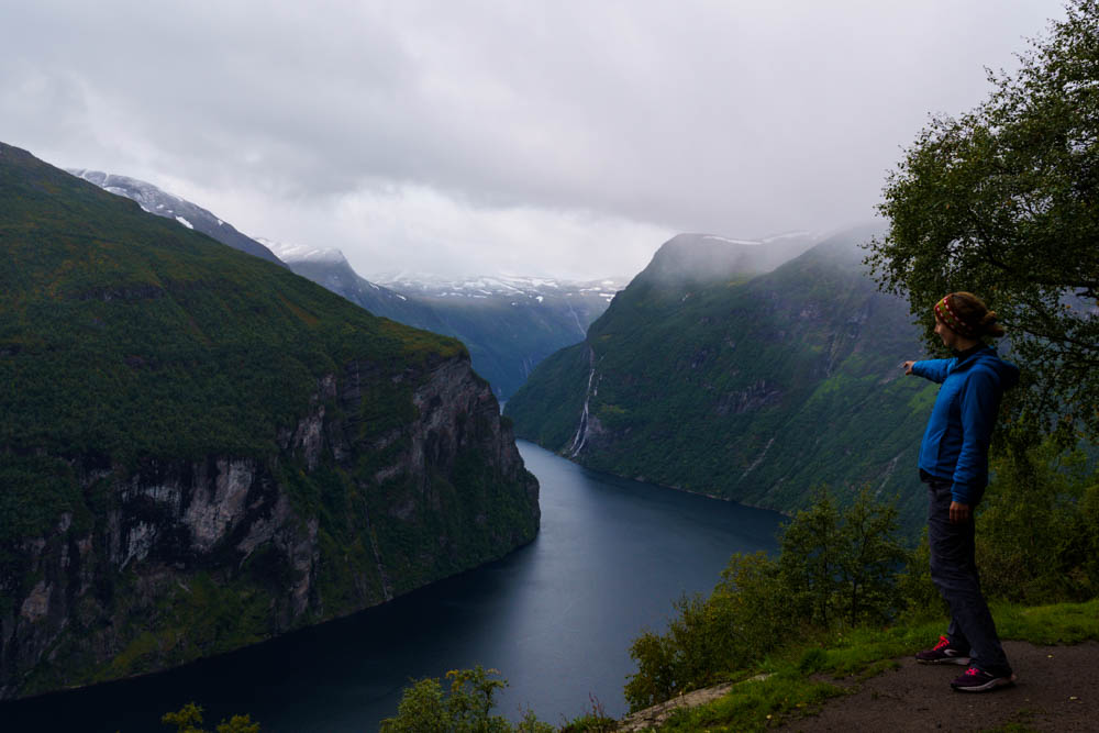 Melanie steht an einem Aussichtspunkt oberhalb des Geiranger Fjord uns zeigt in die Tiefe. Norwegen Bergpässe, Fjorde und Wasserfälle