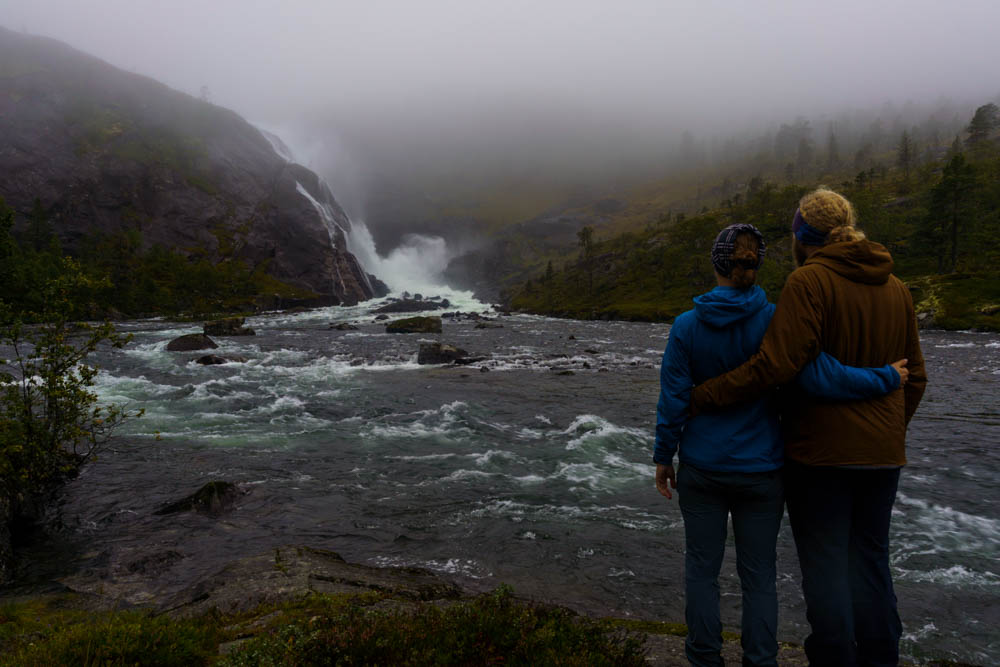 Melanie und Julian stehen mit dem Rücken zur Kamera. Vor ohnen ist ein breiter Fluss welche am Ende von einem Wasserfall gespeist wird. Norwegen Bergpässe, Fjorde und Wasserfälle