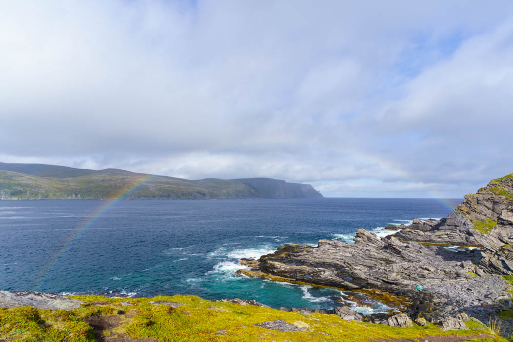 Regenbogen über dem Atlantik beim Kirkeporten nähe Skarsvag. Die dominierenden Farben auf dem gesamten Bild sind blau und türkis im Wasser, eine saftige grüne Wiese im Vordergrund und der Himmel mit seinen Wolken. Fjorde, Berge und Seen, Norwegen