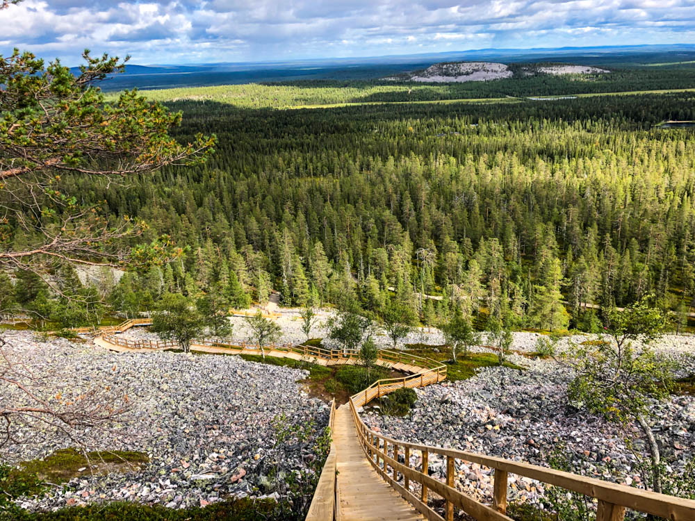Eine lange Holztreppe schlängelt sich vom Gipfel des Luosto Fjell bis ins Tal. Der Horizont scheint nur aus Bäumen und Fjell sowie ein paar Wolken zu bestehen.
