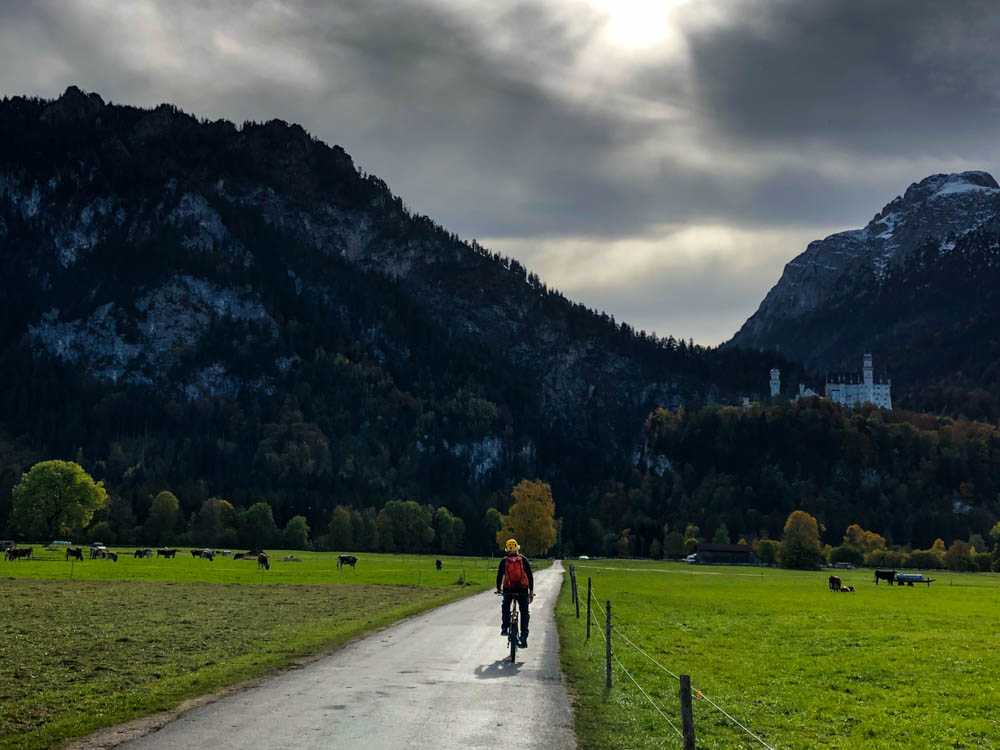 Tour durch die deutschen Alpen - Julian fährt mit dem Fahrrad, dabei ist rechts das Schloss Neuschwanstein, links und direkt vor ihm der Tegelberg zu sehen. Himmel ist wolkig, aber die Sonne kämpft sich schon durch.