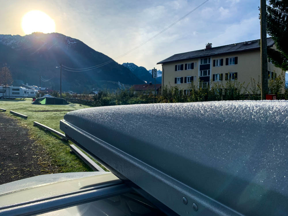 Die Sonne geht über den Gipfeln der Berge von Oberstdorf auf. Der Frost auf der Dachbox ist zu erkennen und glitzert im Sonnenlicht. Tour durch die deutschen Alpen