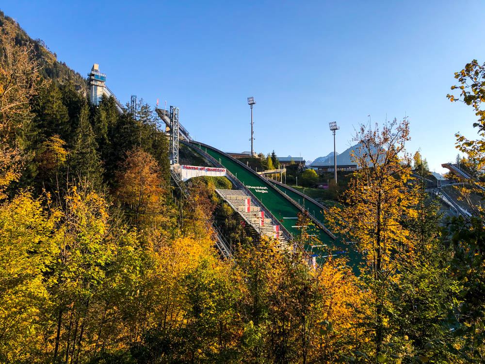 Ein Blick auf das Skisprungstadion in Oberstdorf. Die umliegenden Bäume sind Herbstlich verfärbt und der Himmel blau.
