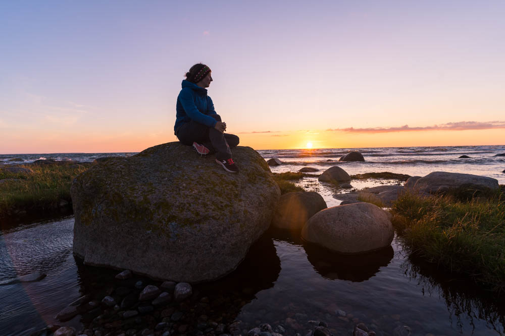 Melanie sitzt auf einem Stein auf Purekkari und im Hintergrund versinkt die Sonne im Meer. Der Himmel ist orange und rot gefärbt.
