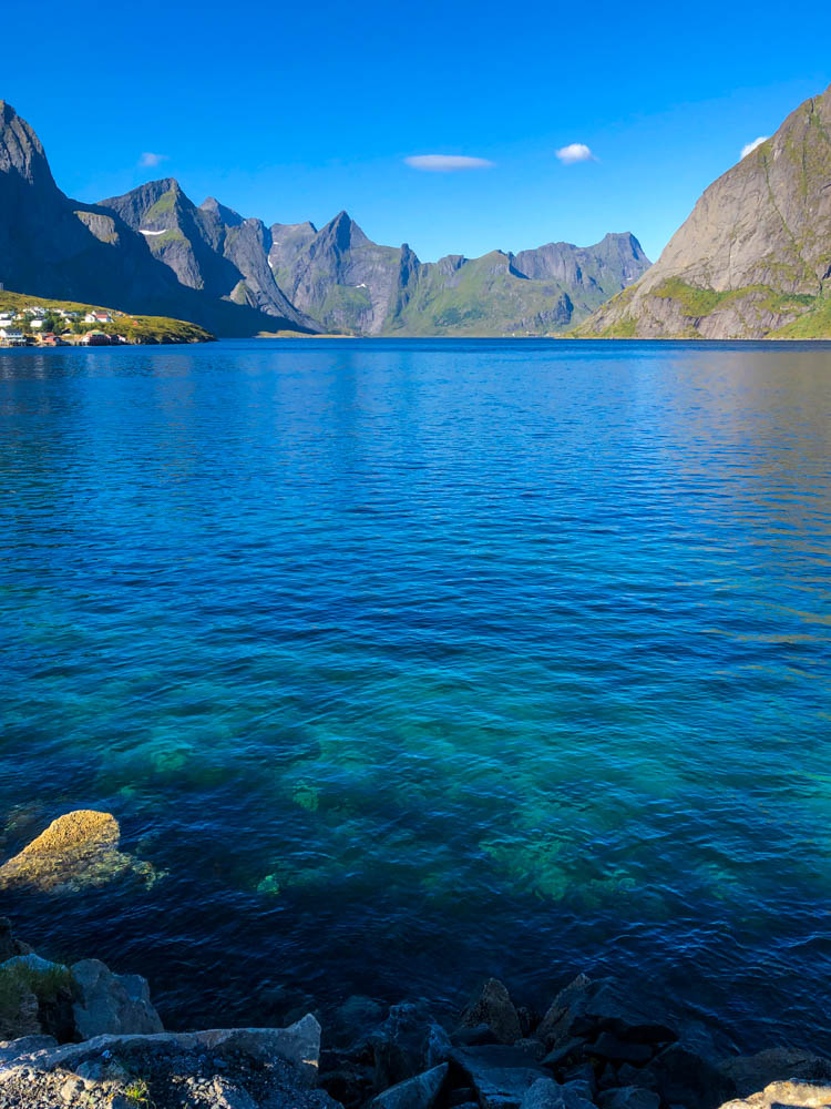 Blick in Reinefjord. Dunkelblaues Wasser im Vordergrund und blauer Himmel über den Bergen am Horizont. Lofoten Alpen mitten im Meer