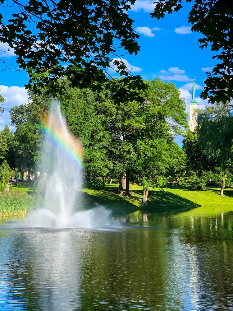 In einem Wasserspiel im Stadtpark von Riga (Lettland) bildet sich ein Regenbogen. Im Hintergrund steht das Freiheitsdenkmal.