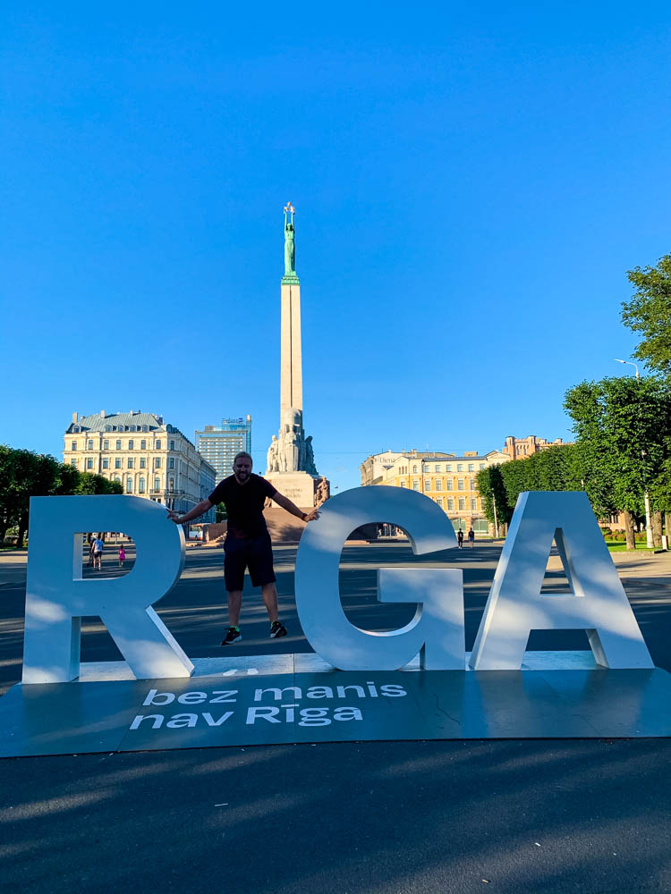 Vor dem Freiheitsdenkmal steht der Stadtname Riga. Julian bildet dabei das "i" und der Himmel ist strahlend blau..