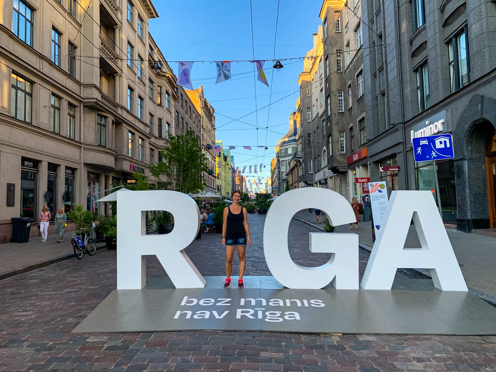 Melanie steht vor der Fußgängerzone in Riga und bildet das "i" des Stadtnamens Riga.