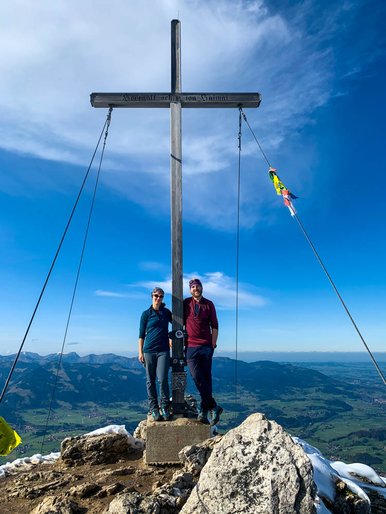 Melanie und Julian stehen am Gipfelkreuz des Rubihorn. Der Himmel ist blau und nur wenige Schleierwolken sind zu sehen. Tour durch die deutschen Alpen
