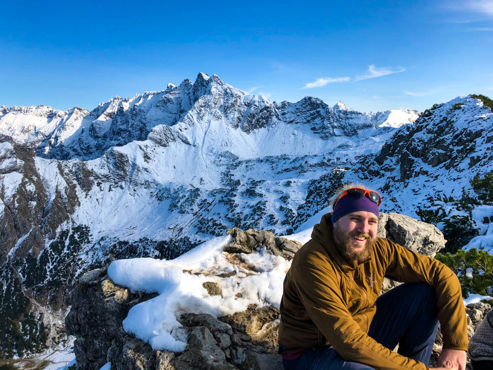 Julian sitz am Gipfel des Rubihorn und lacht in die Kamera. Im Hintergrund sind die schneebedeckten Berge zu sehen. Tour durch die deutschen Alpen