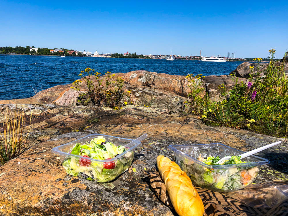 Im Vordergrund stehen zwei Salate und ein Baguette auf einem Stein am Fjell. Im Hintergrund is Helsinki zu sehen. Der Himmel ist blau und wolkenlos. Skandinavien wir kommen