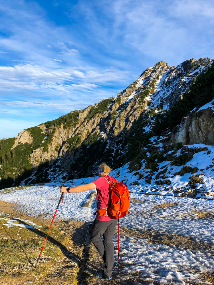 Tour durch die deutschen Alpen - Julian stützt sich auf seine Stöcke. Auf dem Weg liegt eine dünne Schneeschicht. Im Hintergrund sieht man einen Gipfel.