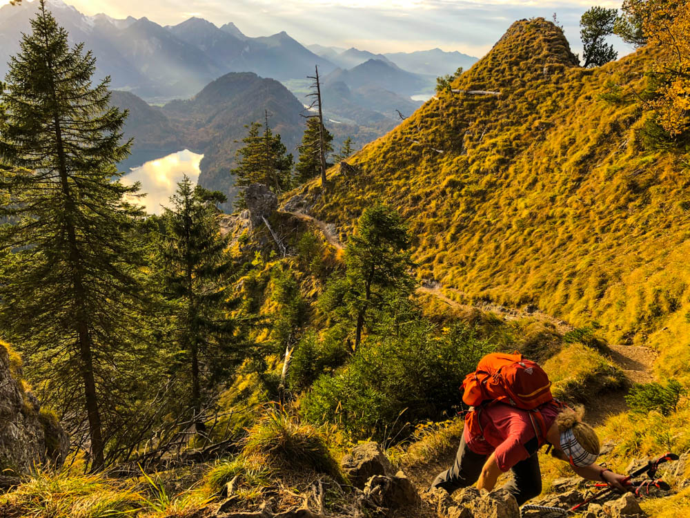 Tour durch die deutschen Alpen - Julian klettert ein Stück rückwärts ab. Der Wanderweg geht steil abwärts. Die Landschaft ist orange und grün gefärbt.