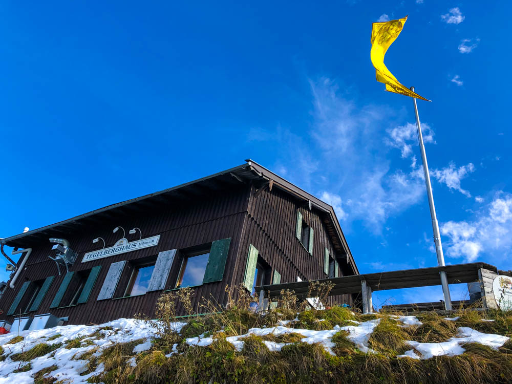 Tour durch die deutschen Alpen - Das Tegelberghaus und eine gelbe Fahne die im Wind weht vor einem strahlend blauen Himmel.