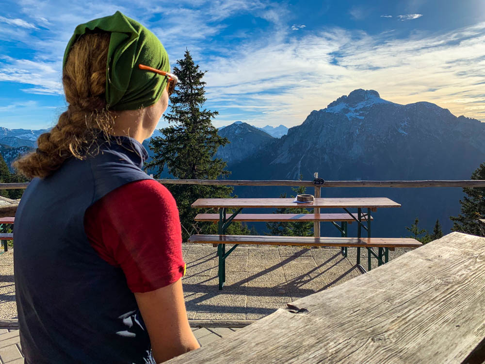 Tour durch die deutschen Alpen - Melanie sitzt an einem Tisch vor dem Tegelberhaus. Im Hintergrund sieht man die Alpen in alle Schönheit.