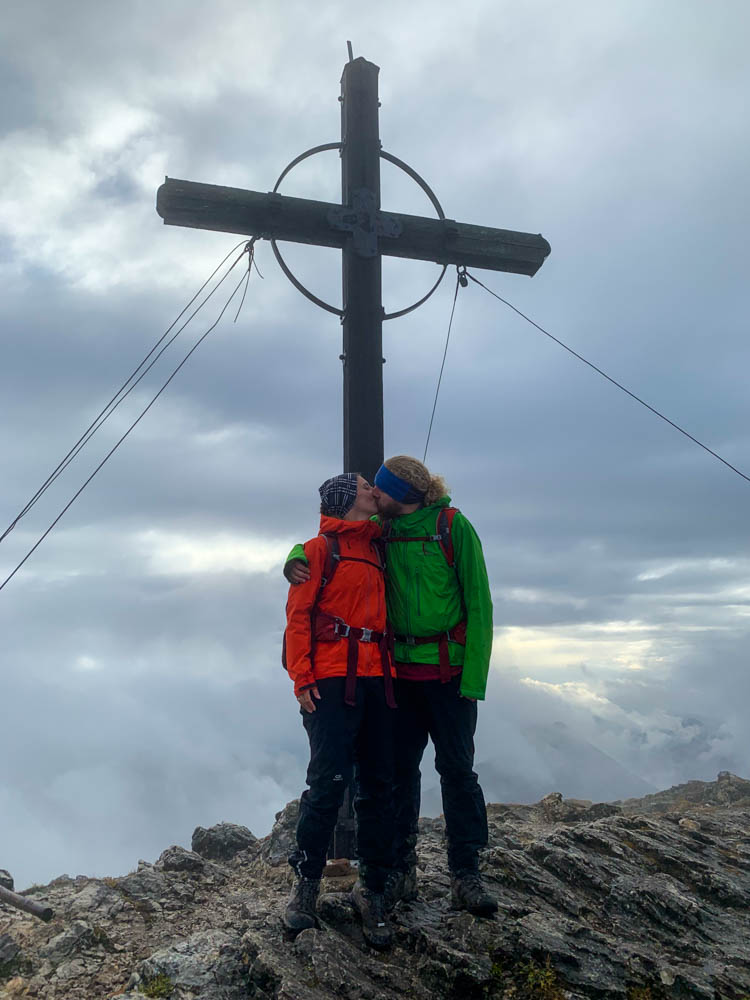 Deutsche Alpen - Melanie und Julian küssen sich unter dem Gipfelkreuz am Roßstein. Die Wolken haben sich verzogen und es hat ein wenig aufgeklart.