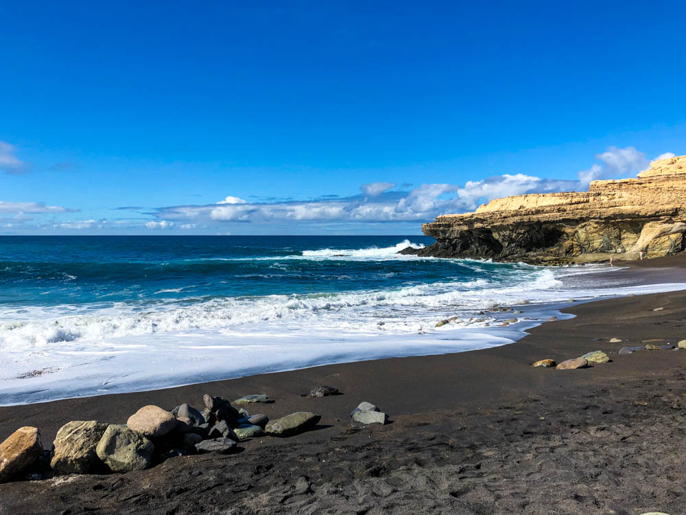 Strand von Ajuy. Der Sand ist dunkel, das Wasser türkisblau und die Felsen im Hintergrund rechts leuchten gelb. Erkundungstouren auf Fuerteventura