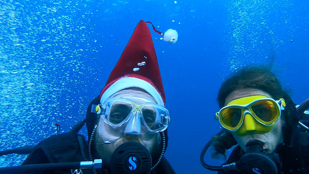 Melanie und Julian machen ein Selfie beim Tauchen. Beide haben einen Atemregler im Mund, die Maske auf und Julian trägt zusätzlich eine Nikolausmütze. Diese steht spitz nach oben da sie durch die Schwerelosigkeit unter Wasser "schwebt". Wassersport im Süden von Fuerteventura