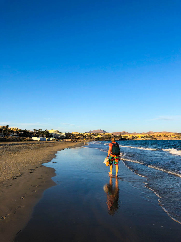 Julian läuft am Strand entlang bepackt mit unseren Einkäufen für unsere Auszeit auf Fuerteventura. Der Himmel ist blau und keine Wolke zu sehen.