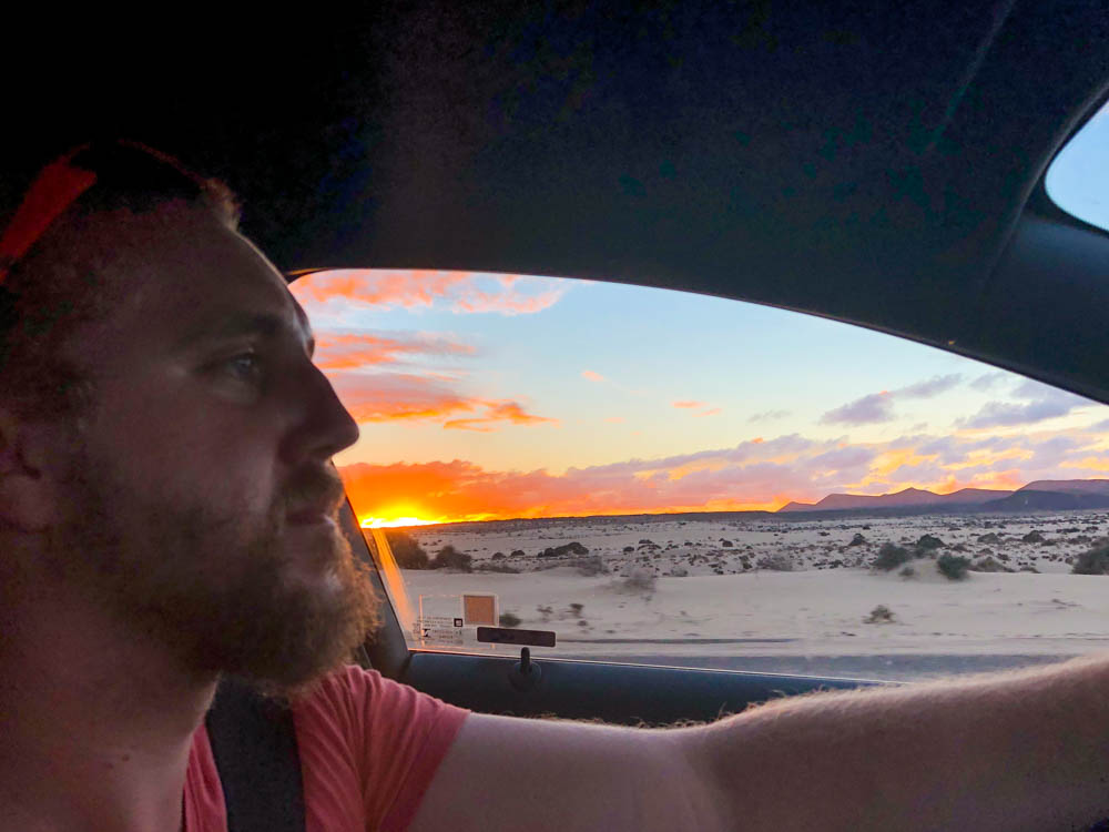 Julian fährt Auto und durch die Fensterscheibe ist der Sonnenuntergang über den Dünen zu sehen.