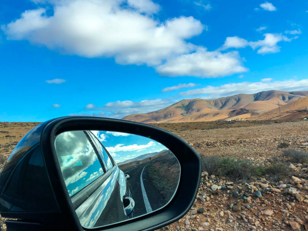 Hügellandschaft Fuerteventura, Fotografiert wurde aus der Fensterscheibe heraus. Der Außenspiegel wurde mit fotografiert und zeigt die Straße. Erkundungstouren auf Fuerteventura