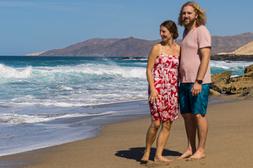 Melanie und Julian stehen an einem Strand, im Hintergrund rollen die Wellen an Land. Der Fokus des Bildes ist auf den Hintergrund gerichtet. Beide sind leicht unscharf. Keiner von beiden schaut in die Kamera, jeder schaut in eine andere Richtung man sieht aber das Lächeln von beiden. Reiseliebe wächst und gedeiht