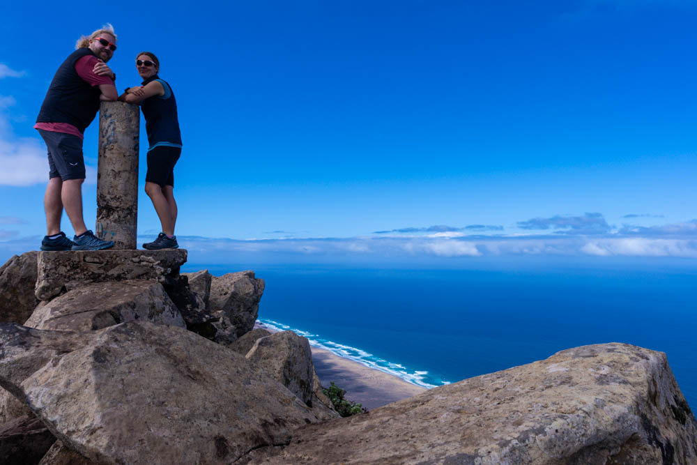 Melanie und Julian stehen am Gipfel Pico de la Zarza und stützen sich auf einer Säule. Im Hintergrund ist der Atlantik und der blaue Himmel zu sehen. Erkundungstouren auf Fuerteventura