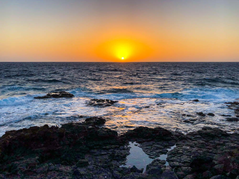 Sonnenaufgang über dem Atlantik. Der Himmel brennt orange während unten die Wellen gegen den Fels branden. Die Sonne ist aufgegangen und die orangene Scheibe schwebt direkt über dem Wasser.