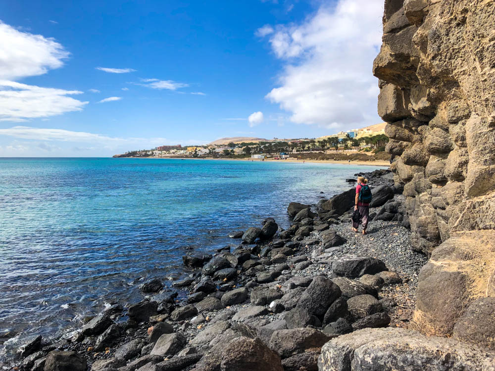 Julian läuft auf einem Steineigen Strandabschnitt in Costa Calma. Recht ragt eine steile Wand aus Lavagestein nach oben. Links ist das türkise Wasser des Atlantik.
