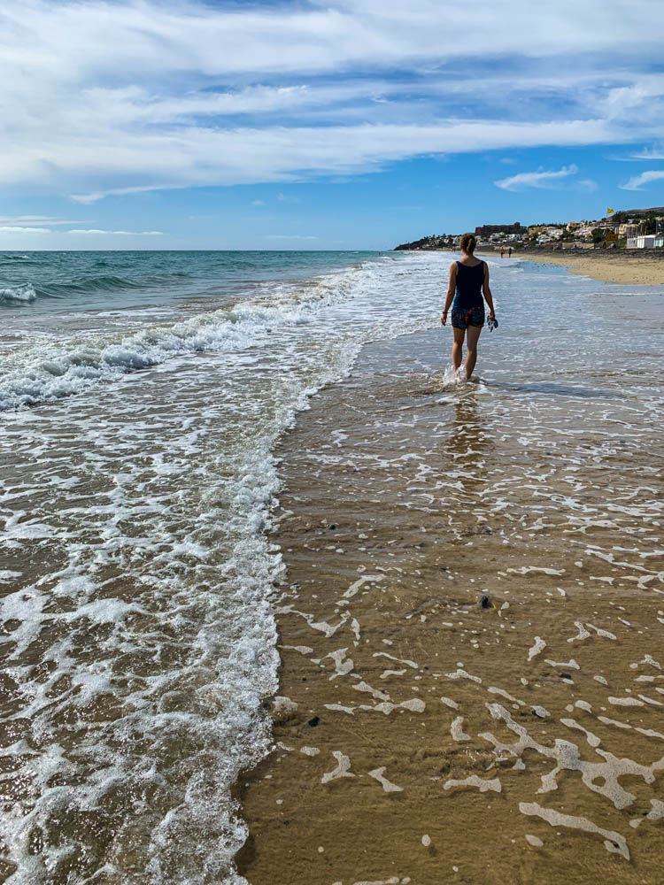 Melanie läuft am Strand von Costa Calma. Das Wasser umspielt ihr Knöchel. Der Himmel ist blau, die perfekte Auszeit auf Fuerteventura
