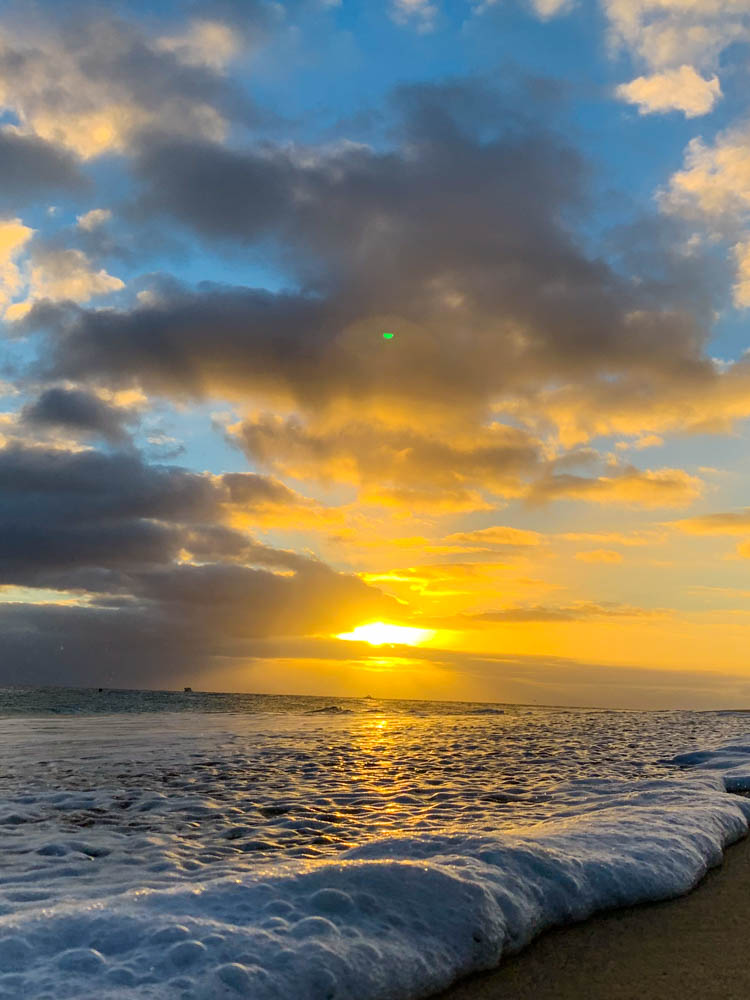 Sonnenuntergang über dem Meer. Die Kameraperspektive ist auf Bodenhöhe. Eine Welle schwappt an den Sandstrand und die Schaumkrone ist direkt vor der Kamera. Der Himmel und die Wolken sind orange gefärbt