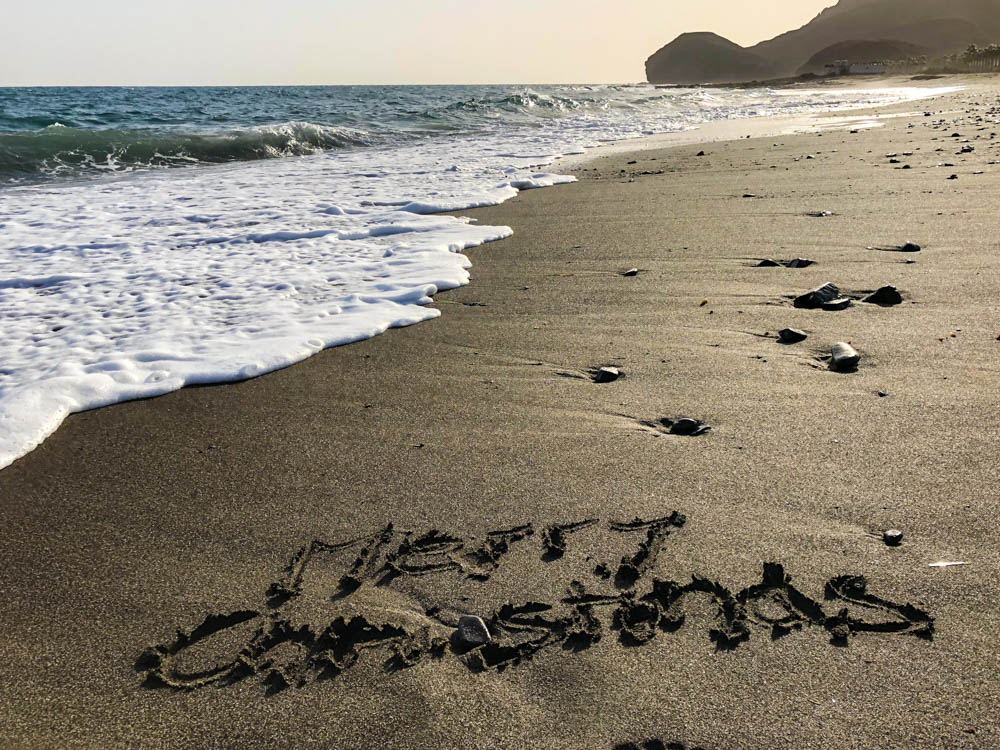 In den Sand am Strand sind die Worte "Merry Christmas" geschrieben. Es ist der Strand, Wellen, welche von links kommen sowie eine bergige Silhouette zu sehen. Weihnachten unter Palmen