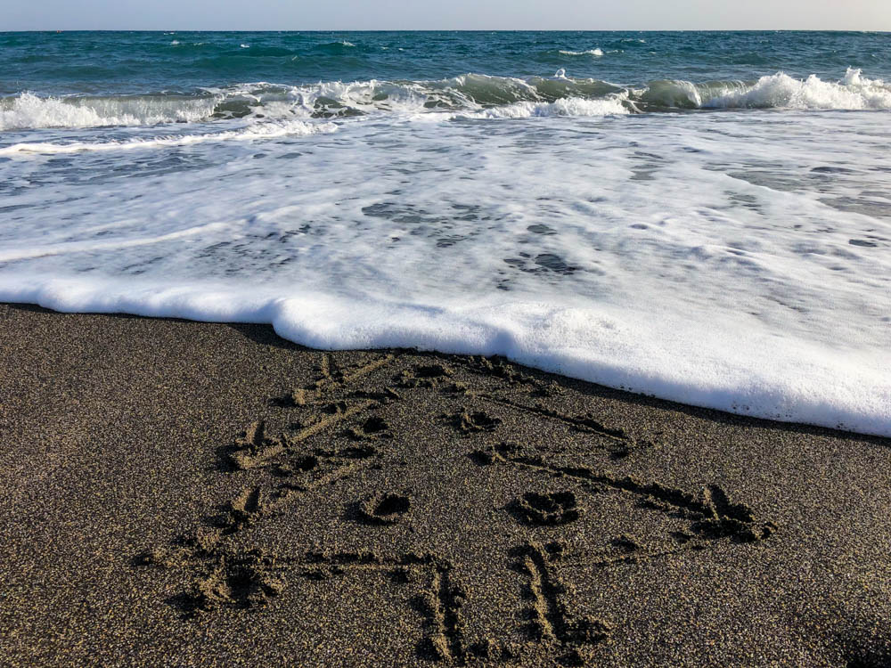 In den Sand am Strand wurde ein Weihnachtsbaum mit Kerzen und Christbaumkugeln gemalt. Im Hintergrund rollen die Wellen herbei und der Weihnachtsbaum wird langsam weggespült.