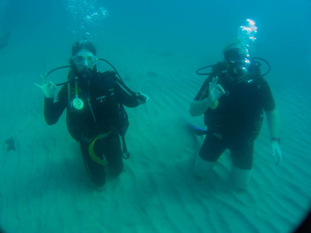 Bild von Melanie und Julian bei ihrem aller ersten Tauchgang. Beide geben das OK Zeichen während sie auf dem sandigen Meeresgrund knien.
