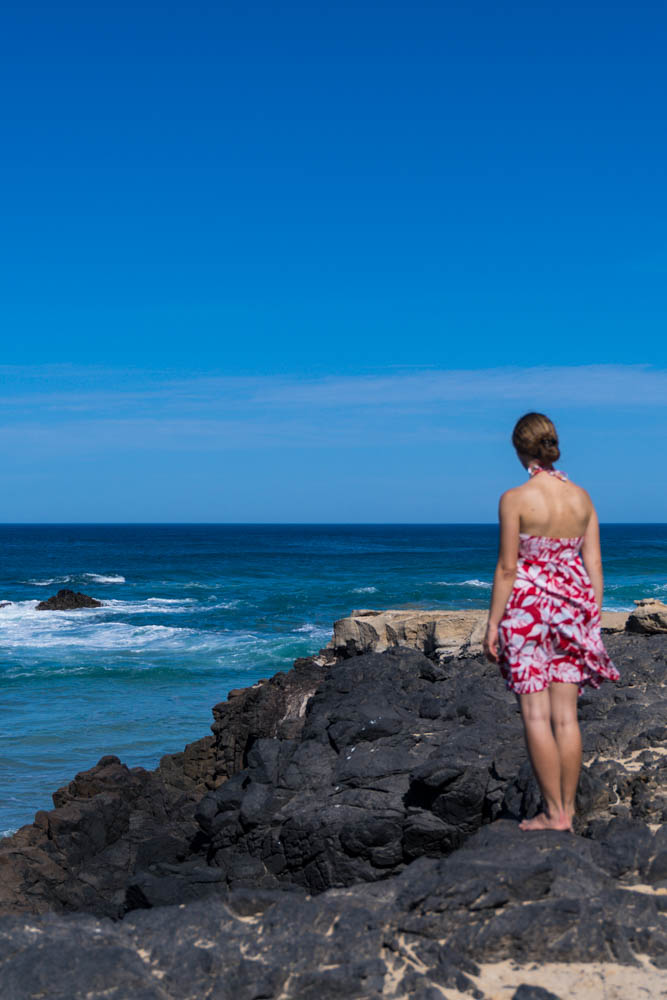 Melanie steht auf einem Felsen an der Westküste. Ihr Sommerkleid weht im Wind. Sie schaut in die Ferne und genießt die Auszeit auf Fuerteventura.