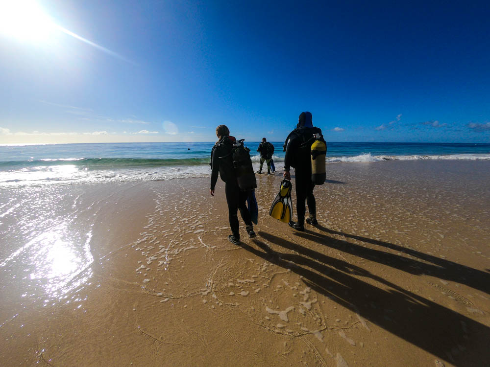 Melanie und Bernd (unser Tauchlehrer) laufen am Strand in Jandia ins Wasser. Beide haben ihre volle Ausrüstung angelegt und spüren die ersten Wellen an den Füßen im flachen Wasser. Wassersport im Süden von Fuerteventura