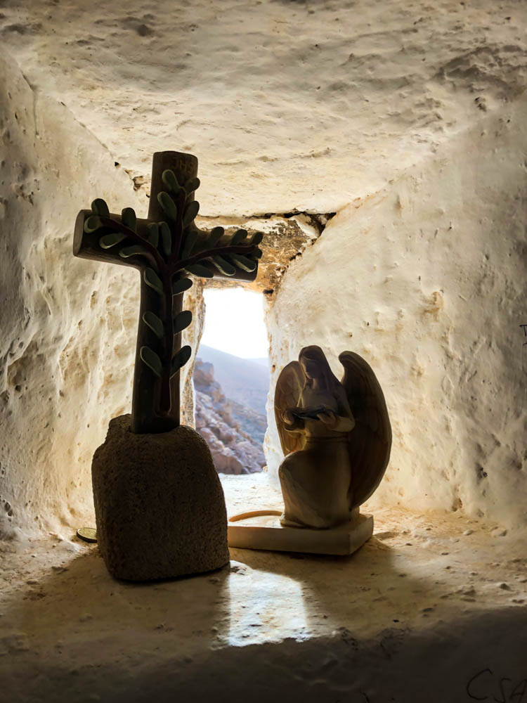 Durch ein kleines Fenster in der Kapelle von Barranco de las Peñitas fällt Sonnenlicht. Davor stehen ein Kreuz sowie eine Engelsstatue welche als dunkle Kontur vor dem Licht erscheinen.