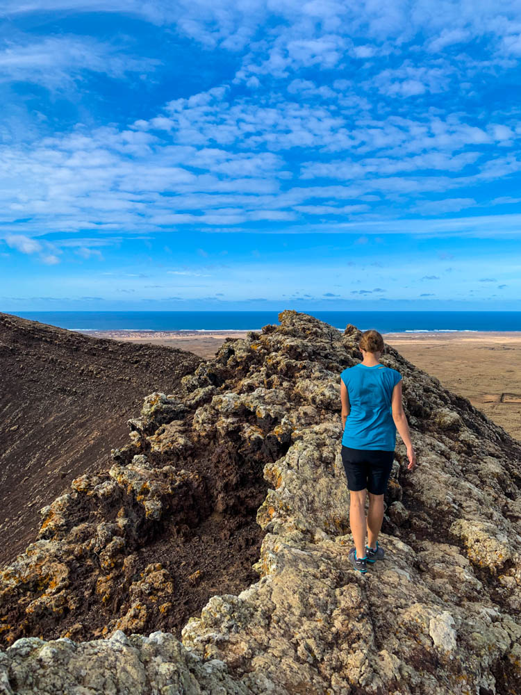 Melanie läuft über den schmalen Grat am Krater von Calderón Hondo. Im Hintergrund sieht man den Atlantik und den blauen Himmel mit kleinen weißen Schleierwolken.