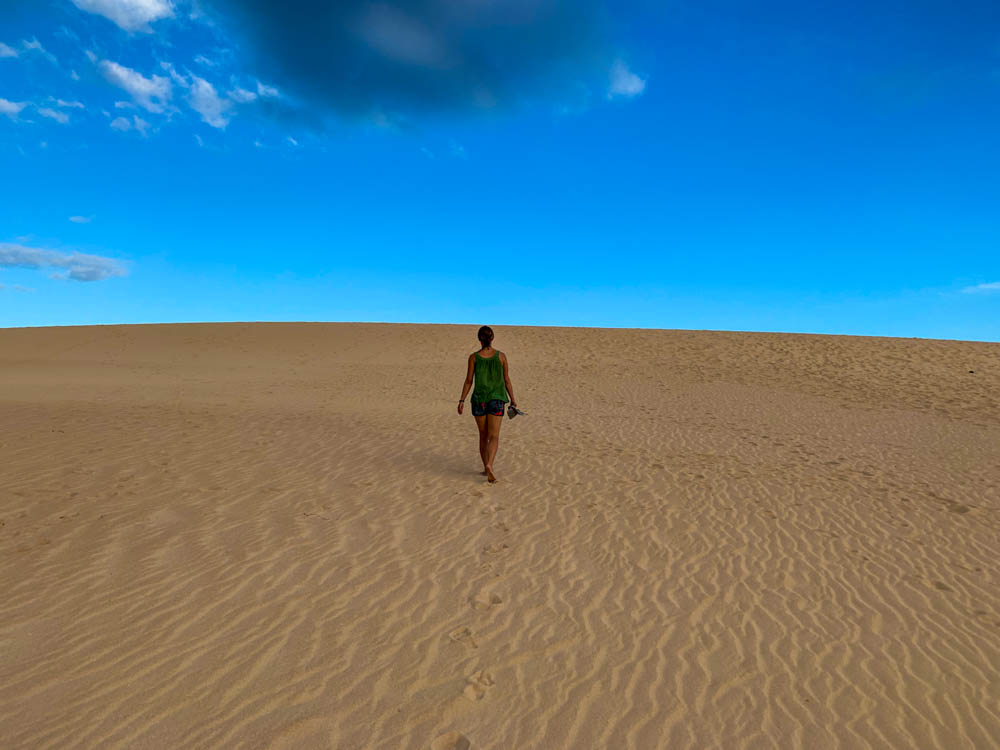 Melanie läuft Barfüßig durch den Sand der Dünen von Corralejo. Der Himmel ist größtenteils blau, nur eine dunkle Wolke ist zu sehen. Die untere hälfte des Bildes besteht nur aus dem San einer Düne über die Melanie läuft.