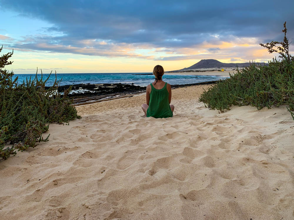 Melanie sitzt im Sand bei den Dünen von Corraleco und schaut auf den Atlantik. Der Himmel ist leicht vom Sonnenuntergang gefärbt. Rechts und Links sind niedrige grüne Büsche, welche dem Bild einen natürlichen Rahmen verleihen.