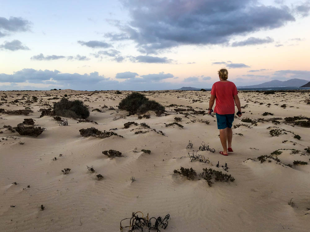 Julian läuft durch die Dünenlandschaft von Corralejo. Es sind niedrige Büsche die auf dem losen Sand wachsen zu sehen. Der Himmel verfärbt sich leicht vom Sonnenuntergang. Fuerteventura, wir bleiben etwas länger