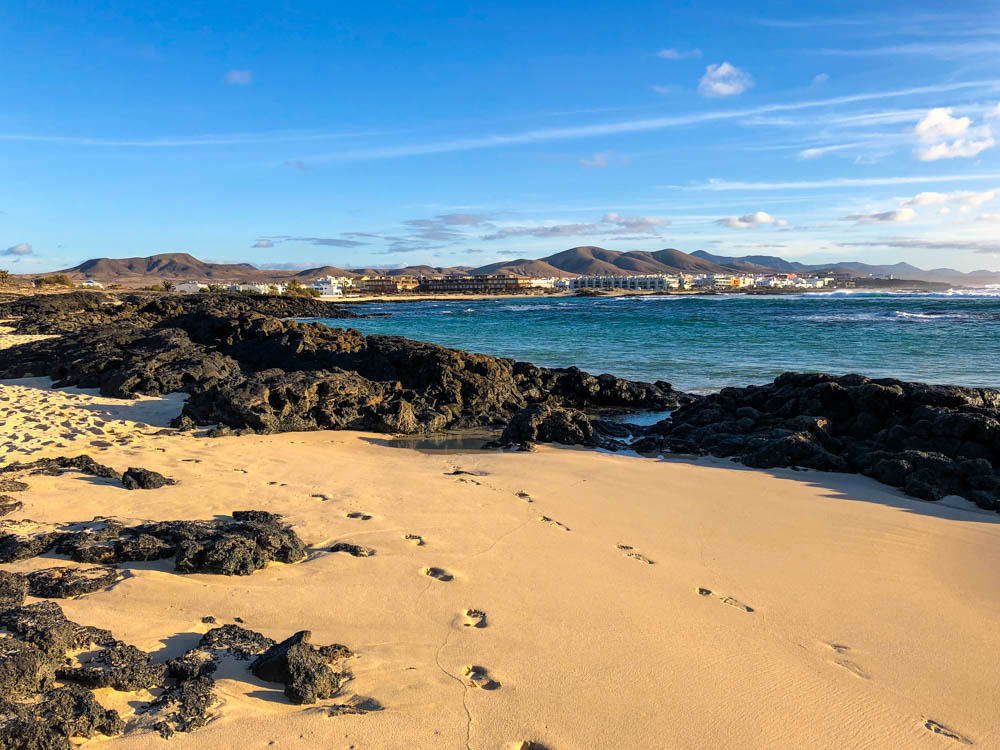 Aufnahme vom Strand im El Cotillo. Es wechseln sich weicher gelber Sand mit dunklem Lavagestein ab. Zwischen dem Strand am unteren Bildrand und dem Ort El Cotillo mit dem blauen Himmel am Horizont liegt eine Bucht mit türkisenem Atlantik.