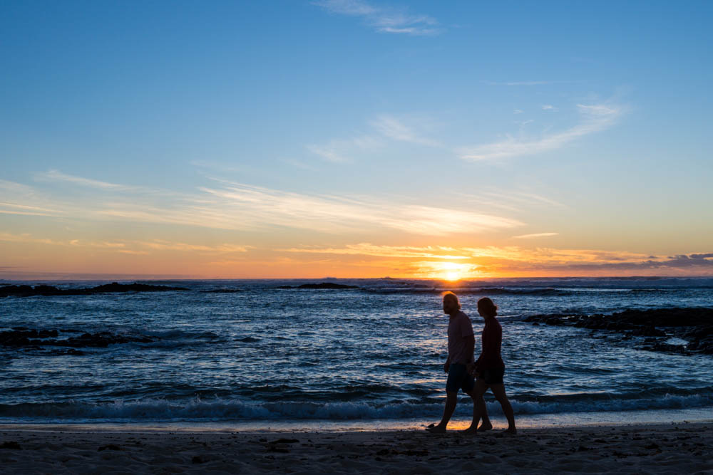 Sonnenuntergang am Strand von El Cotillo. Melanie und Julian laufen Hand in Hand am Wasser entlang. Genau über ihnen versinkt die Sonne in einem Farbenspiel von orange und gelb im Meer.