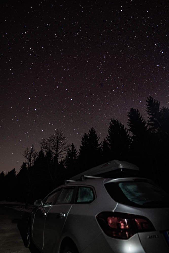Frontaufnahme von Astrarix unter einem gigantischen Sternenhimmel. Das Auto wird von einer Taschenlampe angeleuchtet. Erste Nacht zurück in Deutschland im Camper.