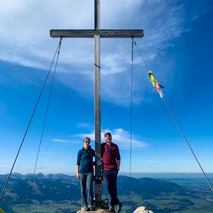 Tour durch die deutschen Alpen. Melanie und Julian stehen neben einem großen Gipfelkreuz.